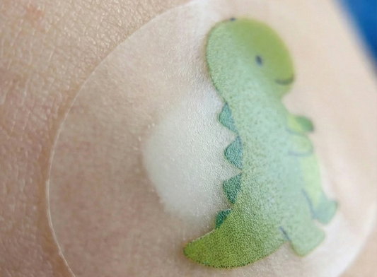 The bandage features a small, green cartoon dinosaur and shows a white, opaque bubble forming in the center, indicating the material is absorbing fluid from a blemish or minor wound.