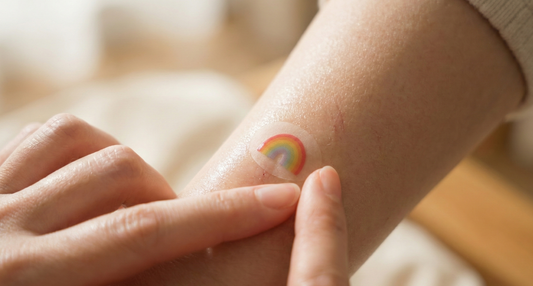 A close-up of a person's arm with a small, circular bandage featuring a colorful rainbow design applied over a minor scratch. Two fingers are gently pressing down on the edges of the bandage.