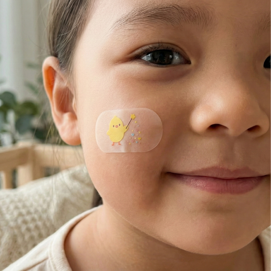 A close-up of a smiling child with a translucent, oval-shaped bandage on their cheek. The bandage features a cute illustration of a small yellow chick holding a star-tipped magic wand with colorful sparkles.